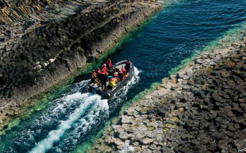 Zodiac boat with Scotland travelers cruises in narrow blue-green water between 2 shores with angular gray rocks.
