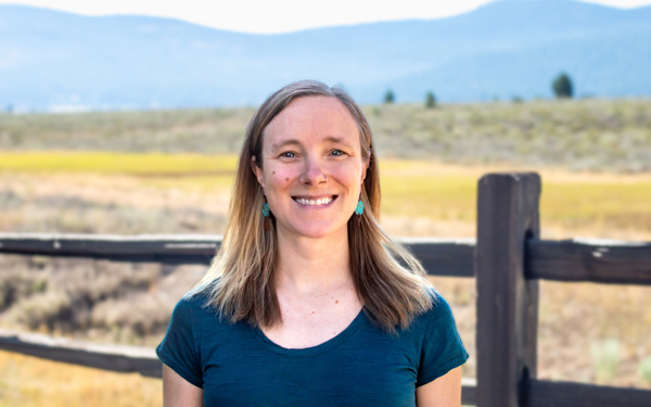 AdventureSmith Content Manager Mary Montalbano with blonde hair & teal shirt stands in front of a wooden fence & golden field.