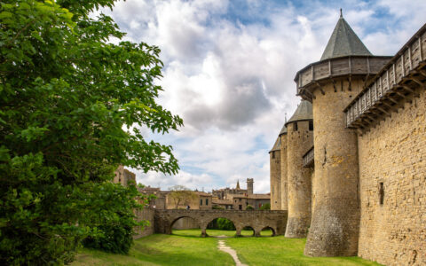 Tall, beige medieval walls with turrets is lined by manicured green grass & deciduous trees under clouds in Carcassone, France.