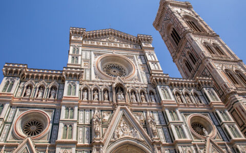 Tall, ornate church with beige & red stone, bell tower, angles & stained-glass circular windows sits under a blue sky in Italy.