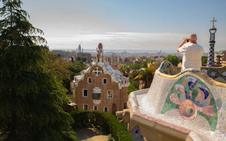Man stands atop colorful mosaic tiled overlook at Park Guell in Spain, photographing the city during a Barcelona to Nice cruise.