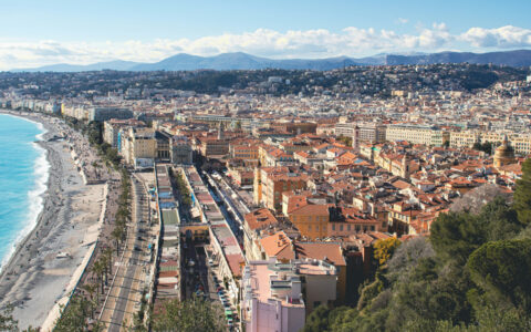Aerial view of Nice, France's teal-sea-lined beach with bright umbrellas backed by terracotta-roofed buildings under a blue sky.