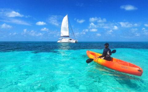 Woman paddles bright orange & yellow kayak in turquoise sea by a white catamaran under sunny skies on a Belize charter cruise.