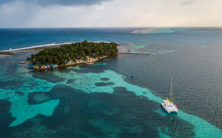 Aerial view of Tobacco Caye, surrounded by turquoise waters, reef & a private catamaran on the Belize Sailing Adventure charter cruise.