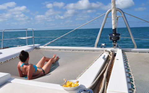 Woman in a bikini lounges on the trampoline netting at the front of her private Belize catamaran charter on a sunny day.