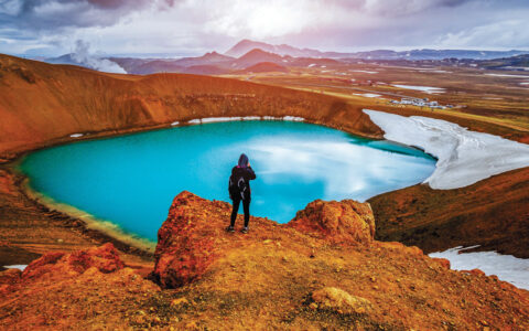 Hiker overlooks geothermal valley with turquoise glacier melt lake & golden-orange hills under cloudy sky by Myvatn Lake, Iceland.