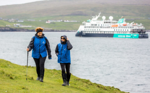 Two women in black pants & blue coats walk seaside bright green grass as blue, white & teal small ship sits in harbor behind.