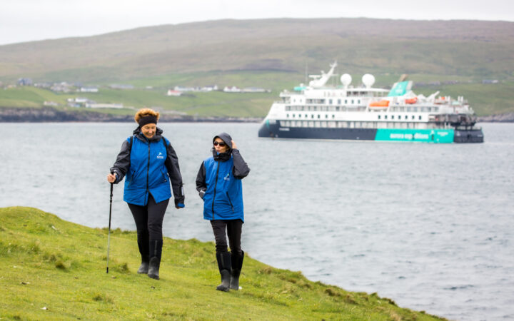 Two women in black pants & blue coats walk seaside bright green grass as blue, white & teal small ship sits in harbor behind.