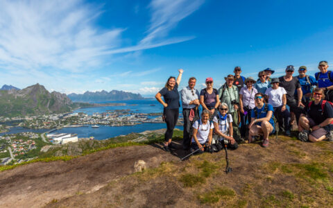 Small group of hikers smile at the summit overlooking a seaside town backed by tall mountains under a blue sky in Norway.