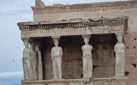 Stone pillars of women hold up the side of an ancient beige building on a cloudy day in Athens, Greece.
