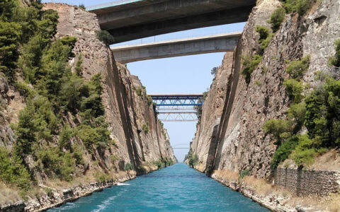 Narrow Corinth Canal with tall beige stone walls, teal water & various bridges on a sunny day in Greece.