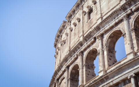 Looking up to top of Rome Italy's ancient colosseum with tall stone pillars creating open-air windows under a blue sky.