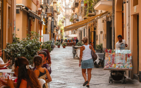 Woman in jean skirt & white tank top walks past snack vendor on cobblestone street as people dine al fresco in Italy.