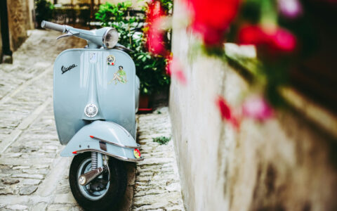 Light blue vespa sits parked beside beige stone wall with hanging red flowers, seen on the Godfather cruise in Italy & Malta.