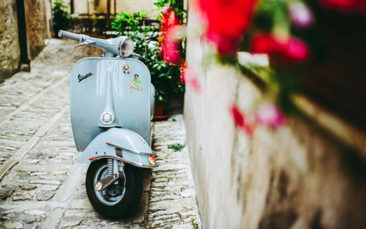 Light blue vespa sits parked beside beige stone wall with hanging red flowers, seen on the Godfather cruise in Italy & Malta.