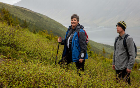 Woman hiker in blue jacket smiles as man in gray shirt looks uphill on a hike in green shrubs along Iceland's Westfjords & North Coast.