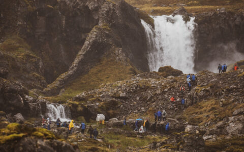 Travelers on Iceland's Westfjords & North Coast cruise hike over tundra & boulders near large waterfall in misty conditions.