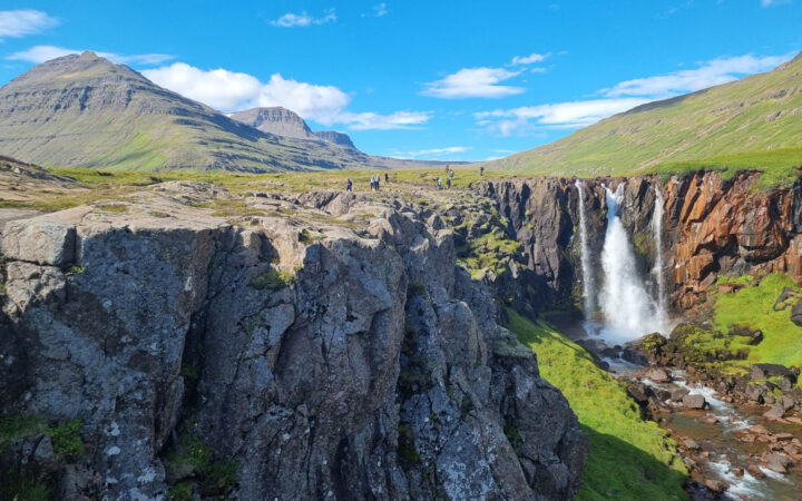 Hikers walk back from large cliffs overlooking gushing waterfall under sunny skies & among green grass in Iceland.