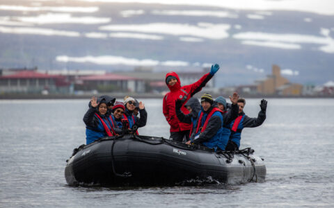 Zodiac boat with waving Iceland's Westfjords & North Coast cruise guests moves through calm water under cloudy skies.