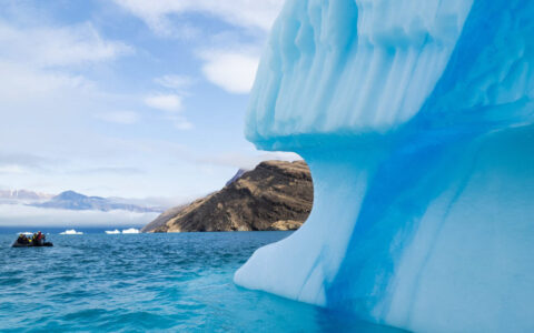 A giant blue iceberg sits on the water in the foreground with a peek of rocky land and a zodiac in the water behind it.