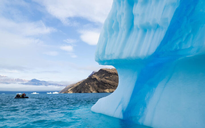 A large blue iceberg floats in the foreground with a zodiac cruising behind it in blue water