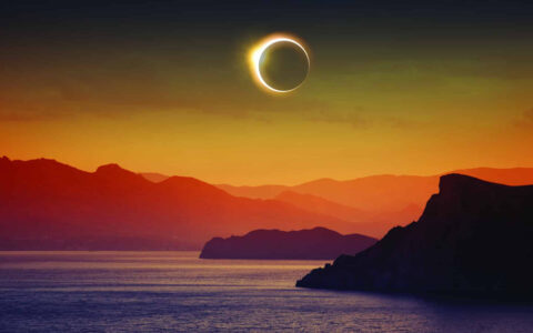 Full solar eclipse with moon blocking sun over arctic landscape of mountains & sea in orange, red, purple & gold.