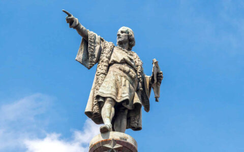 Statue of christopher columbus pointing as a monument to him, infront of a blue sky and light clouds