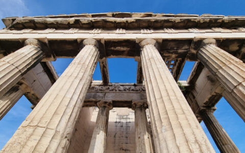 Looking up tall stone pillars holding an open, carved stone roof under a blue sky at the Acropolis in Athens, Greece.