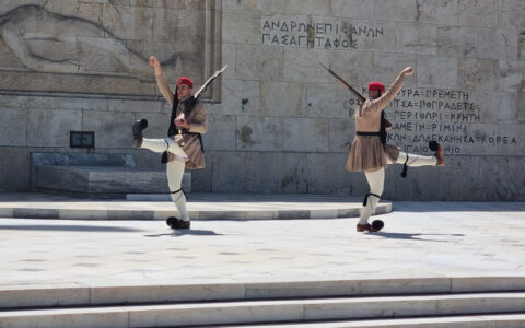 2 armed Greek guards in traditional garb wave arms & legs in a ceremonial changing of the guard atop a beige stone plaza.