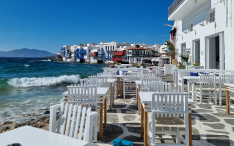 Empty whitewashed tables, chairs & restaurants sit seaside on a sunny day with turquoise waves crashing in Mykonos, Greece.