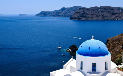 Aerial view of blue-domed, white-washed clifftop building overlooking blue sea on a sunny day in Santorini, Greece.