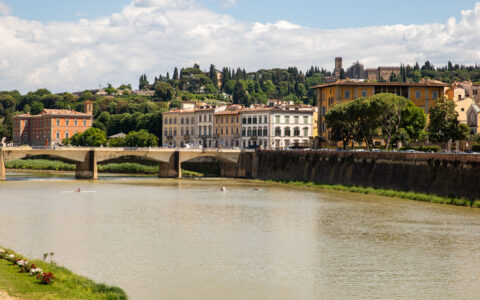 Wide, calm, brown Arno River in Florence, Italy with brick retaining walls & long bridge beside mansions & trees in the sun.