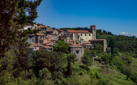 Terracotta-roofed stone buildings in beige & gray sit atop a lush green hillside under blue skies in Cinque Terre, Italy.