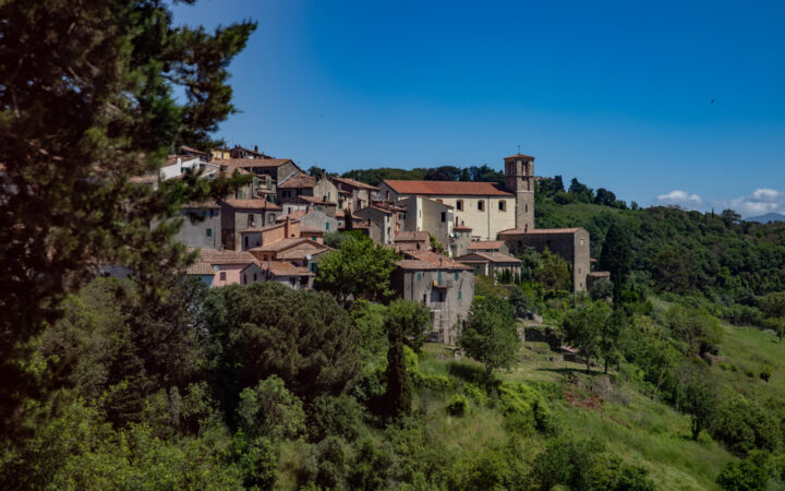 Terracotta-roofed stone buildings in beige & gray sit atop a lush green hillside under blue skies in Cinque Terre, Italy.