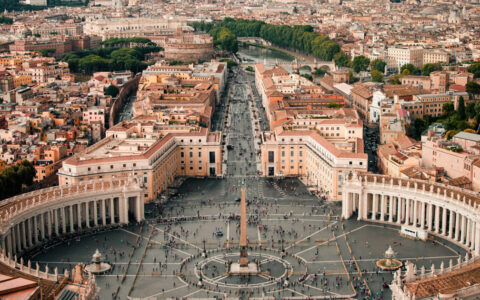 Aerial view of the Vatican in Rome with semicircular buildings around a pillar statue, visitors walking & a nearby river.