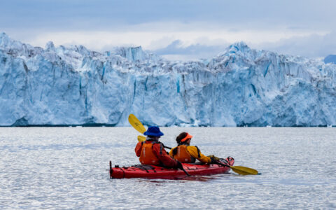 Tandem kayakers in red boat paddle calm water by large blue glacier under cloudy Arctic skies on the Northern Lights Explorer.