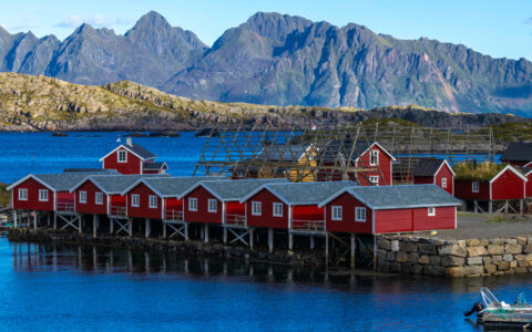A row of side-by-side red houses on stilts sits over calm blue sea backed by striking brown peaks in Lofoten, Norway.