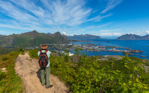 Male hiker in wide-brimmed tan hat walks a dirt trail among green flora, looking down on a small port town backed by mountains.