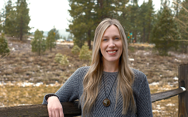 AdventureSmith Olivia Williams standing against a wooden fence next to some pine trees smiling on a cloudy day