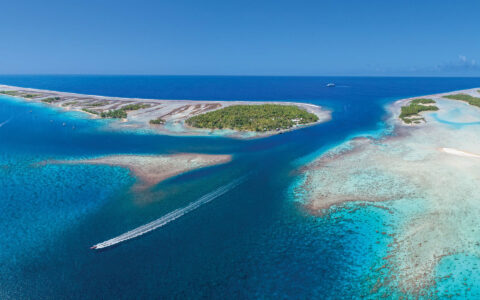 Aerial view of Zodiac zooming past tropical islands & bright coral reefs on a Society & Tuamotu Islands small ship cruise.