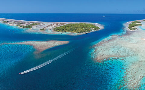 Aerial view of Zodiac zooming past tropical islands & bright coral reefs on a Society & Tuamotu Islands small ship cruise.