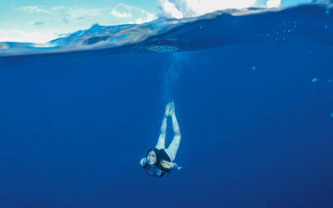 Woman dives below the surface in dark blue water under a sunny sky during a Society & Tuamotu Islands small ship cruise.
