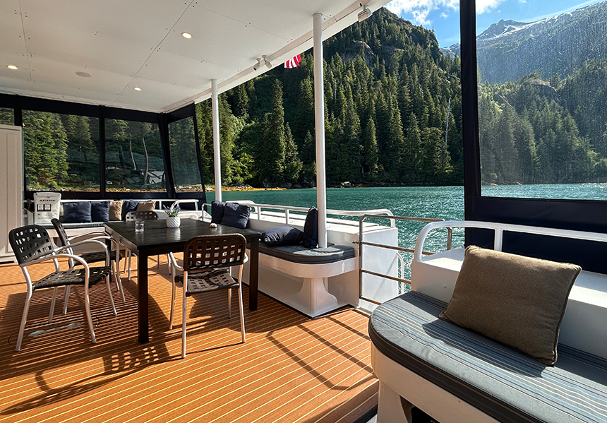 Aft deck of Safari Quest with table and chairs, cushioned seats and pillows surrounded by clear plastic panels for views of Alaska