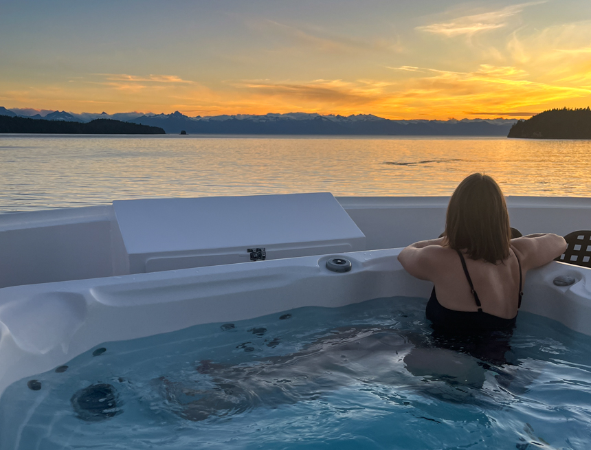 Female traveler in jacuzzi looking out over a quiet bay in Alaska as the sun sets a light orange over the mountains at the horizon.