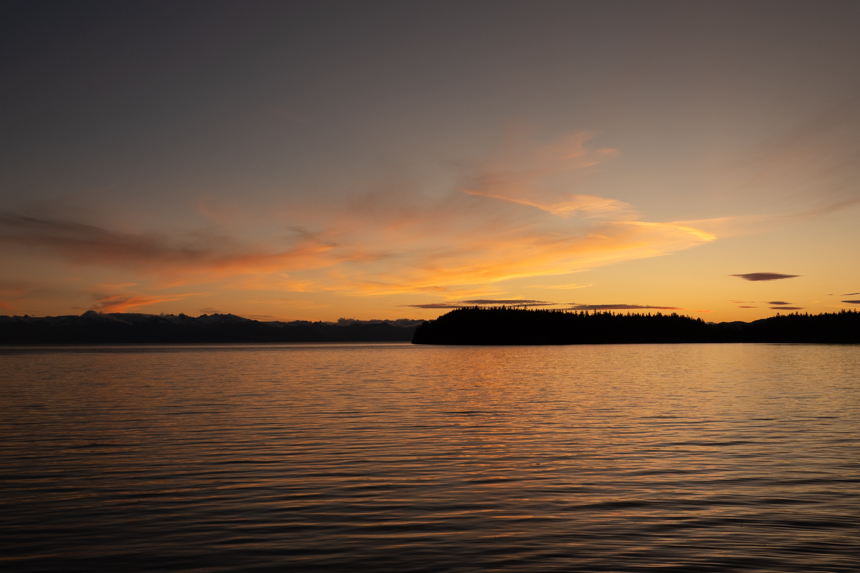 Orange sunset over the water in Alaska with dark silhouette of mountain and forest on the horizon.
