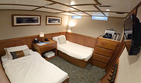 Navigator C4 cabin aboard Safari Quest. Two twin beds with all white bedding, end table between, tv on wall, two framed photos, drawers, binoculars, water bottles and lamp.
