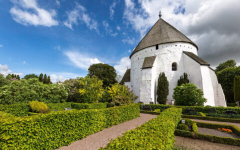 Tall white castle like building stands over a lush green garden on a blue cloudy day