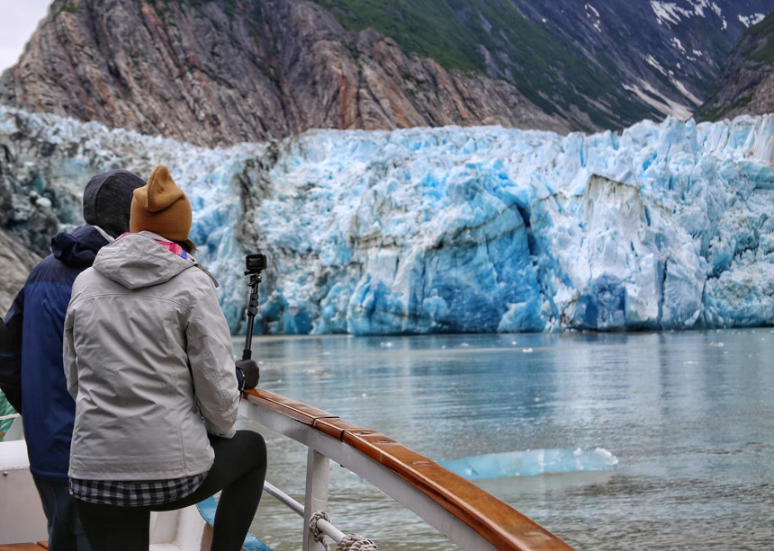Two travelers stand on the bow of a small ship looking out toward a blue glacier