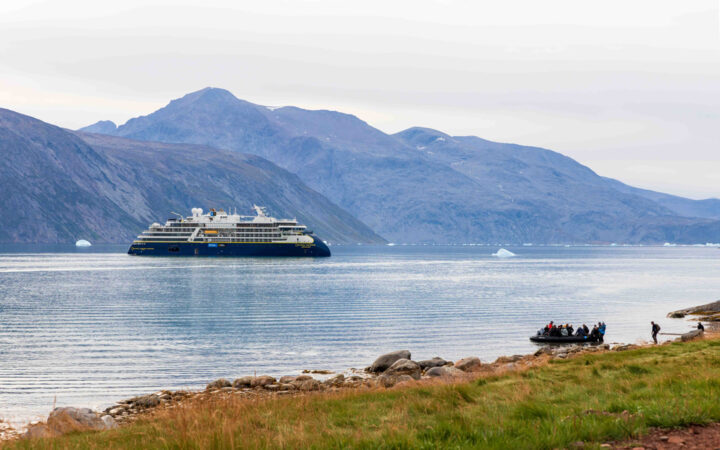 Small ship with dark blue hull & white upper decks sits offshore from grassy coast where Zodiac of Greenland travelers are arriving.