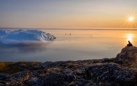 South Greenland Exploration cruise traveler sits alone on top of rocky cliff overlooking calm sea & snoy icebergs at sunset.